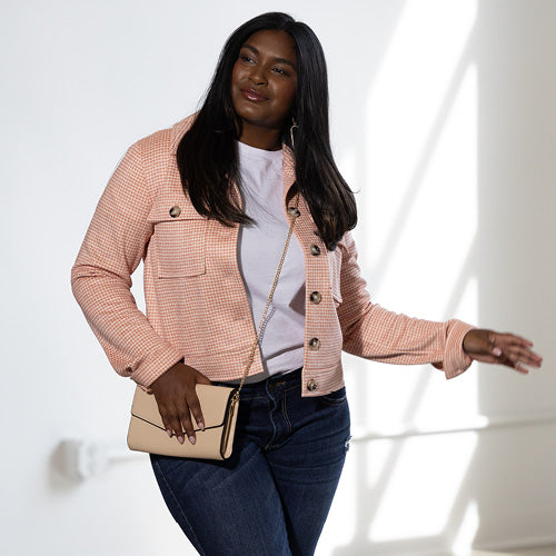 A fashionable woman with long dark hair poses in a peach-colored tweed jacket, white top, and dark denim jeans, accessorized with a neutral crossbody purse. She smiles while looking off to the side in a softly lit room